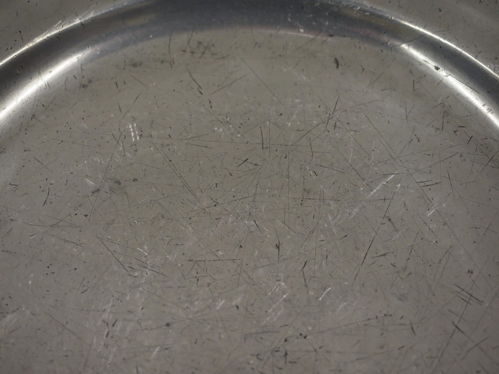 A circular antique pewter charger plate featuring a moulded wavy border and light surface knife marks. View V