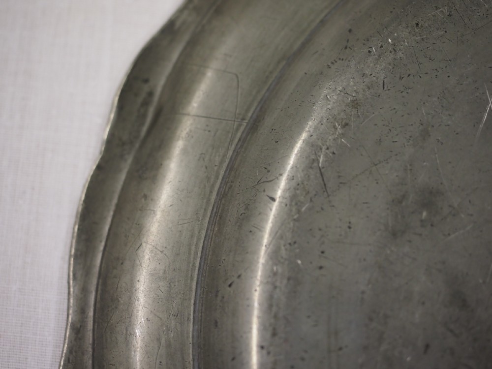 A circular antique pewter charger plate featuring a moulded wavy border and light surface knife marks. View VI