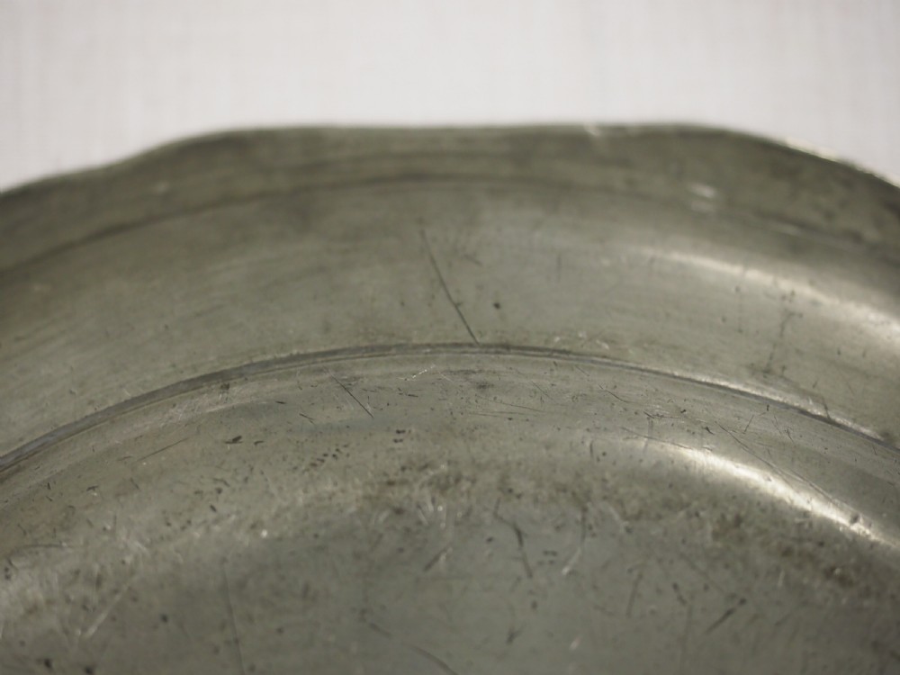 A circular antique pewter charger plate featuring a moulded wavy border and light surface knife marks. View VII