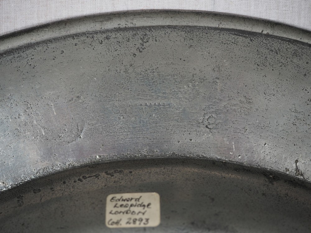 A circular antique English pewter dish featuring a wide rim with multiple reeded edge mouldings and maker touchmarks visible on the lower brim. View VII