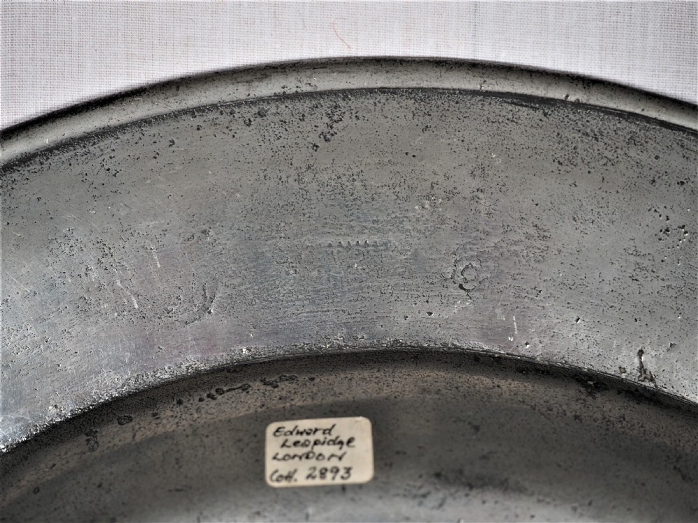 A circular antique English pewter dish featuring a wide rim with multiple reeded edge mouldings and maker touchmarks visible on the lower brim. View VIII