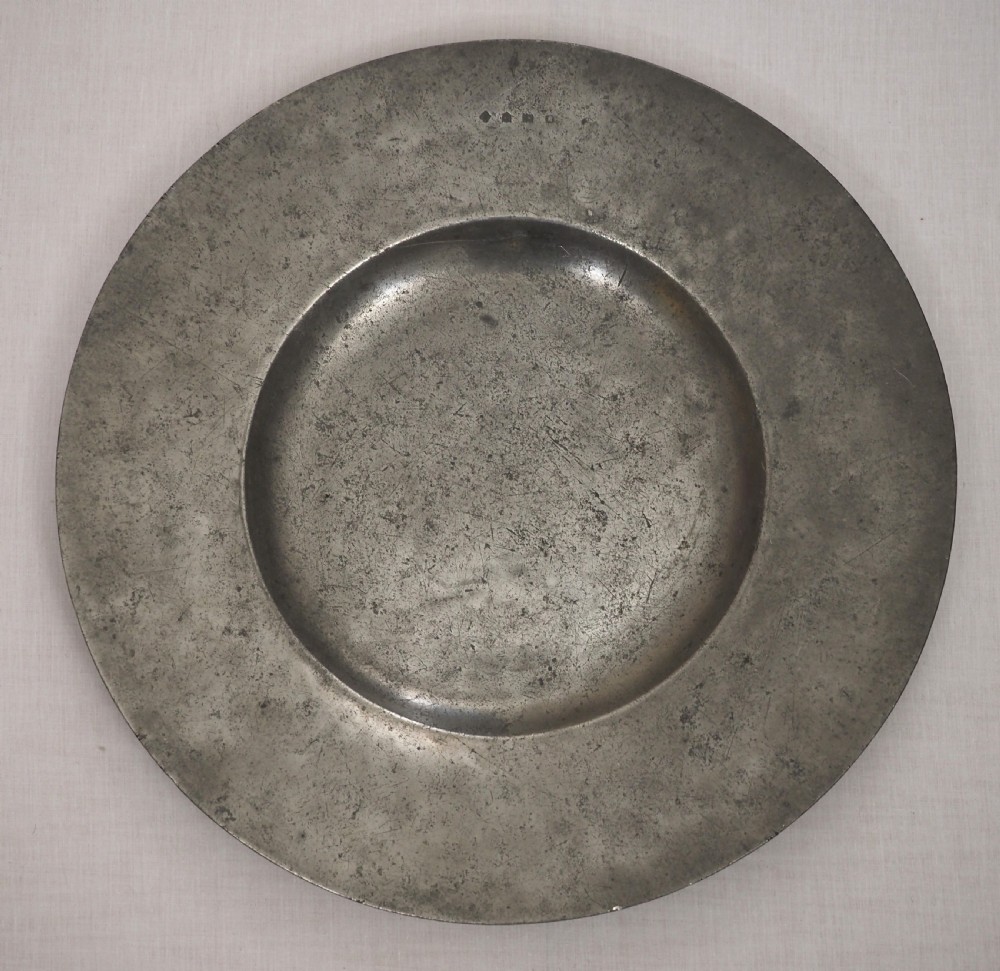 A circular antique pewter charger plate featuring a wide flat rim stamped with hallmarks surrounding a shallow central well.
