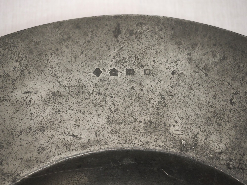 A circular antique pewter charger plate featuring a wide flat rim stamped with hallmarks surrounding a shallow central well. View III