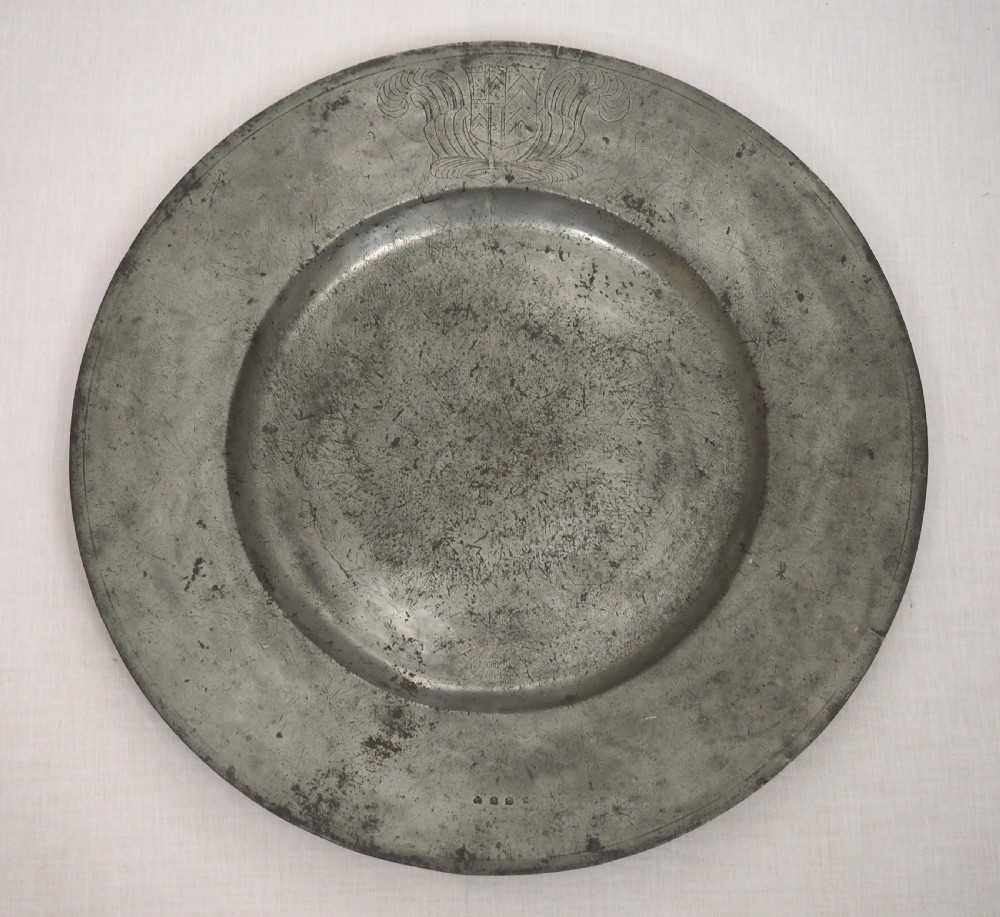 A circular 17th-century pewter charger with a broad rim, featuring an engraved heraldic crest at the top and visible touchmarks at the bottom.