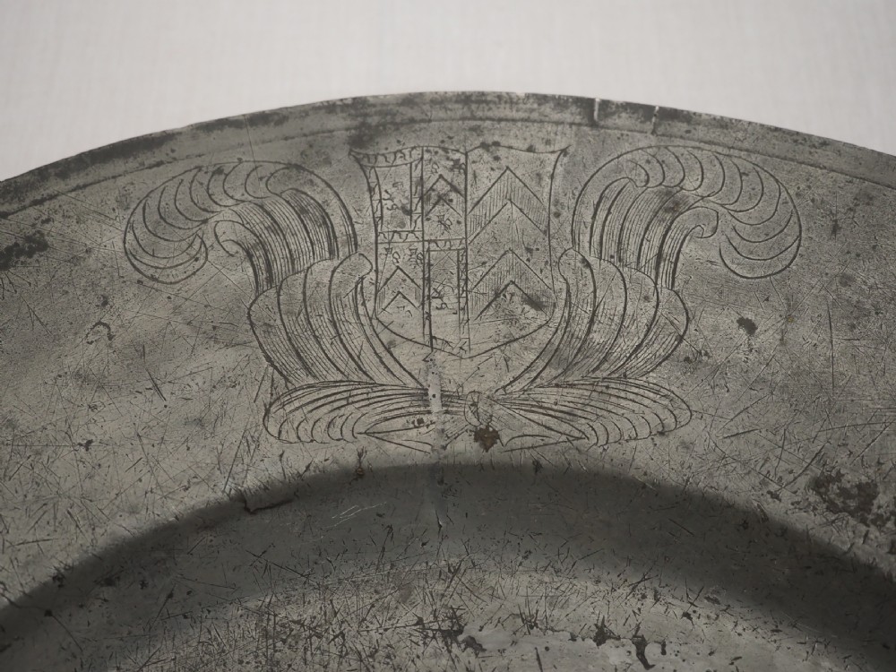 A circular 17th-century pewter charger with a broad rim, featuring an engraved heraldic crest at the top and visible touchmarks at the bottom. View III