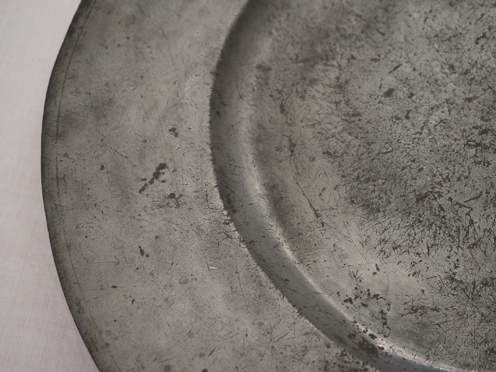 A circular 17th-century pewter charger with a broad rim, featuring an engraved heraldic crest at the top and visible touchmarks at the bottom. View IV
