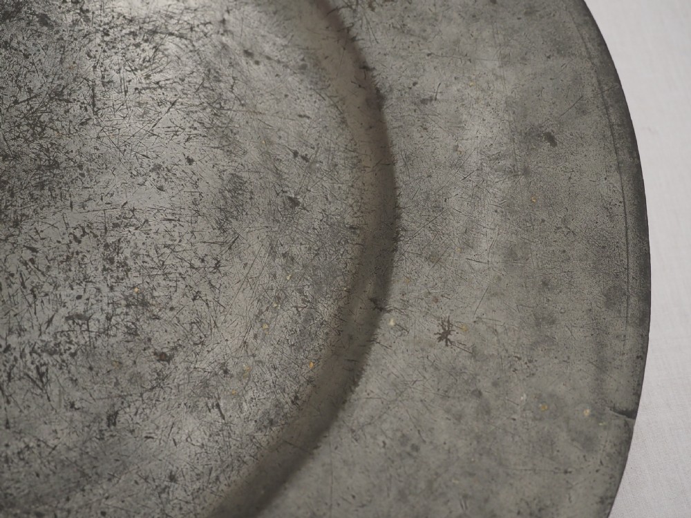 A circular 17th-century pewter charger with a broad rim, featuring an engraved heraldic crest at the top and visible touchmarks at the bottom. View V