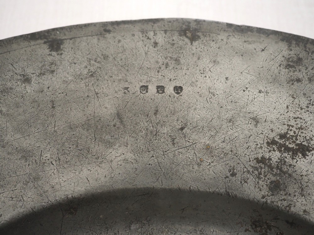 A circular 17th-century pewter charger with a broad rim, featuring an engraved heraldic crest at the top and visible touchmarks at the bottom. View VI