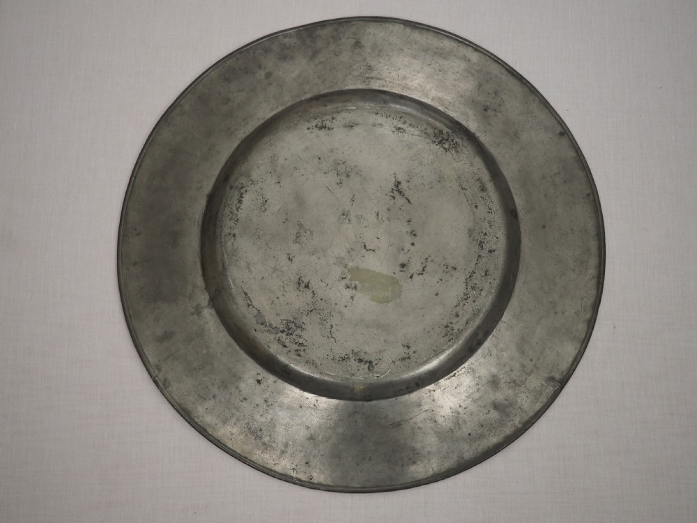 A circular 17th-century pewter charger with a broad rim, featuring an engraved heraldic crest at the top and visible touchmarks at the bottom. View VIII