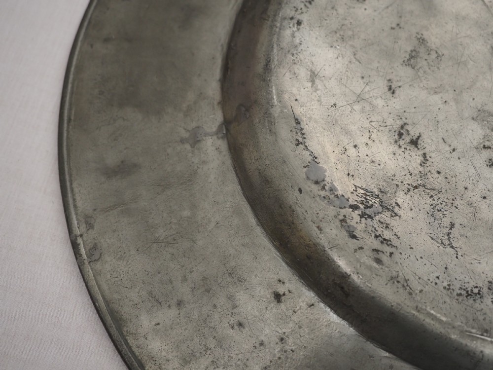 A circular 17th-century pewter charger with a broad rim, featuring an engraved heraldic crest at the top and visible touchmarks at the bottom. View IX