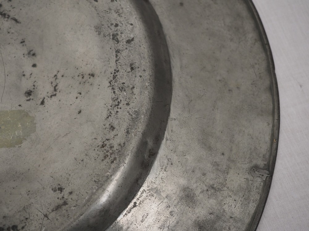 A circular 17th-century pewter charger with a broad rim, featuring an engraved heraldic crest at the top and visible touchmarks at the bottom. View X