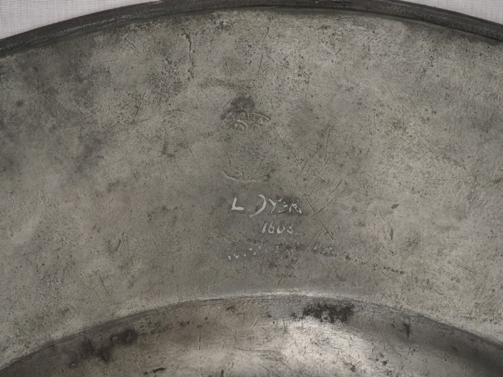 A circular 17th-century pewter charger with a broad rim, featuring an engraved heraldic crest at the top and visible touchmarks at the bottom. View XI