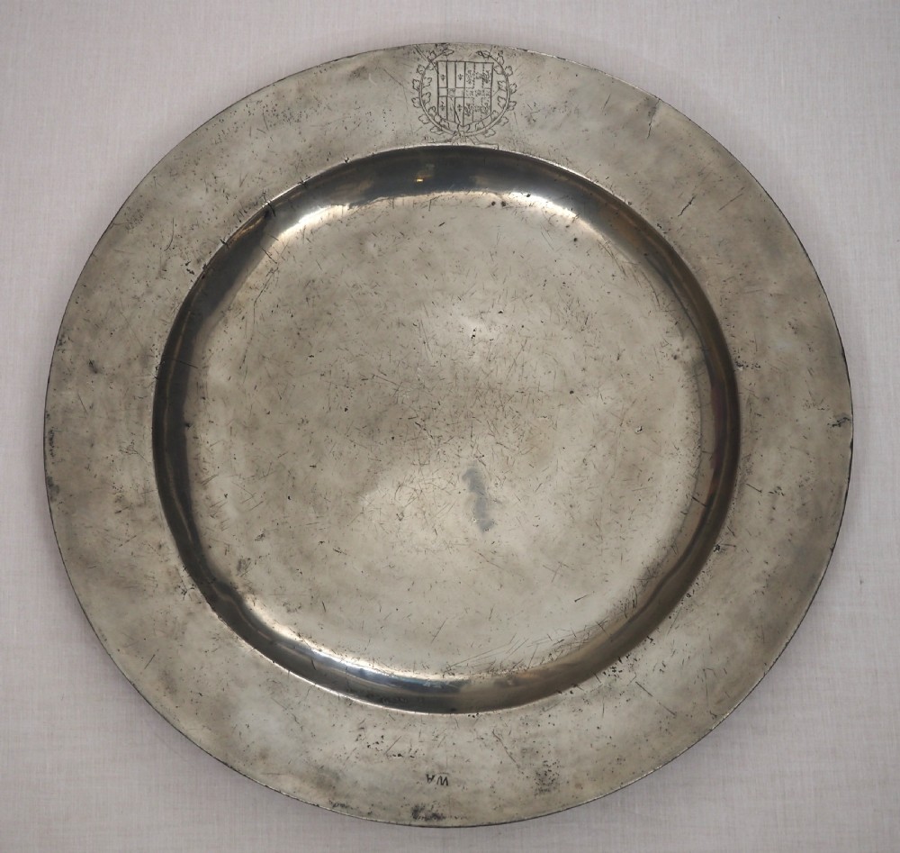 A circular seventeenth-century pewter charger plate with a plain rim featuring an engraved crest and stamped initials.