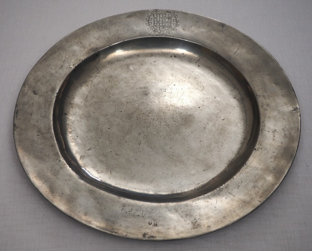 A circular seventeenth-century pewter charger plate with a plain rim featuring an engraved crest and stamped initials. View II
