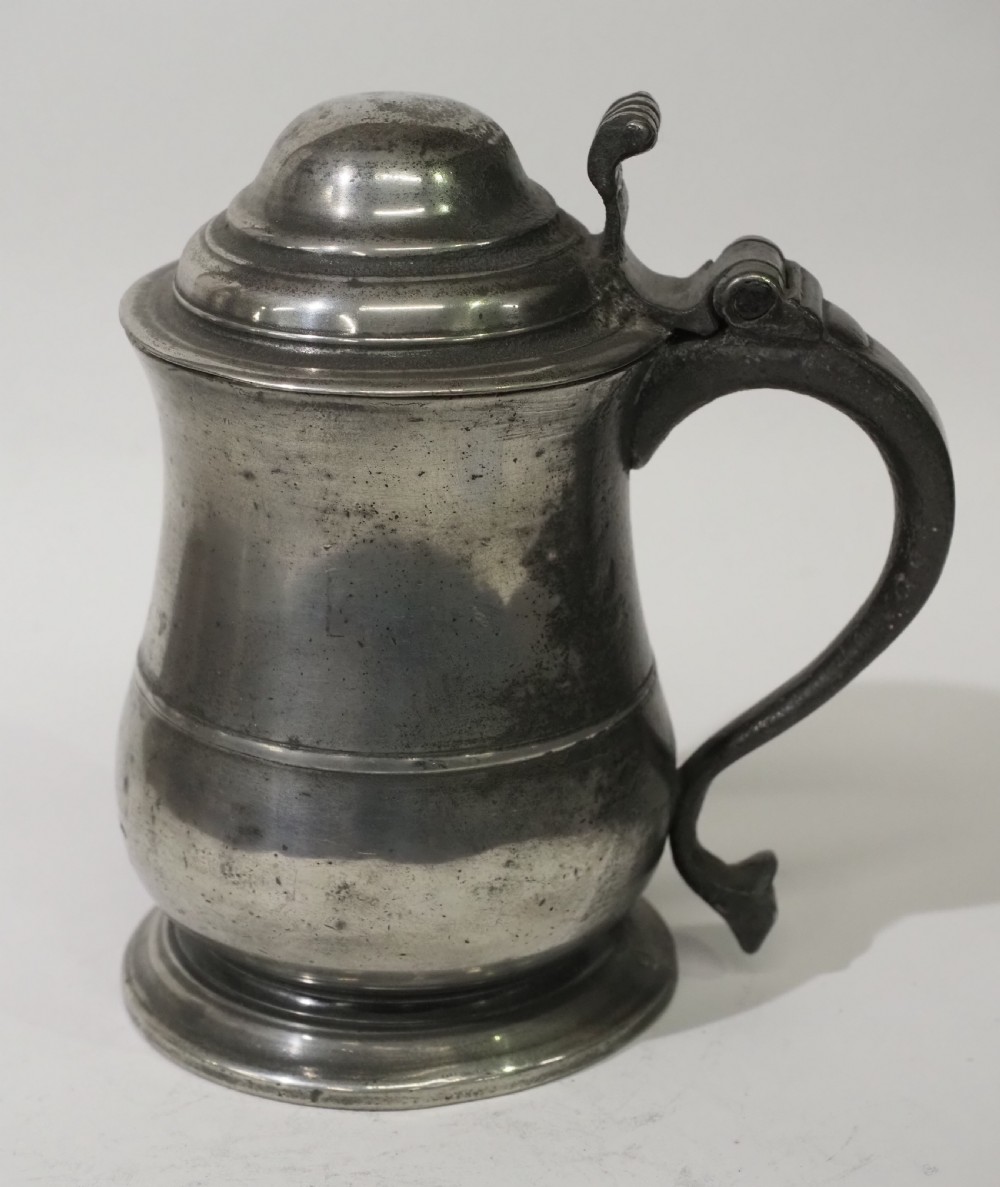 An 18th-century pewter pint tankard featuring a hinged dome lid, a curved scroll handle with a thumbpiece, and a moulded central band on a circular footed base.
