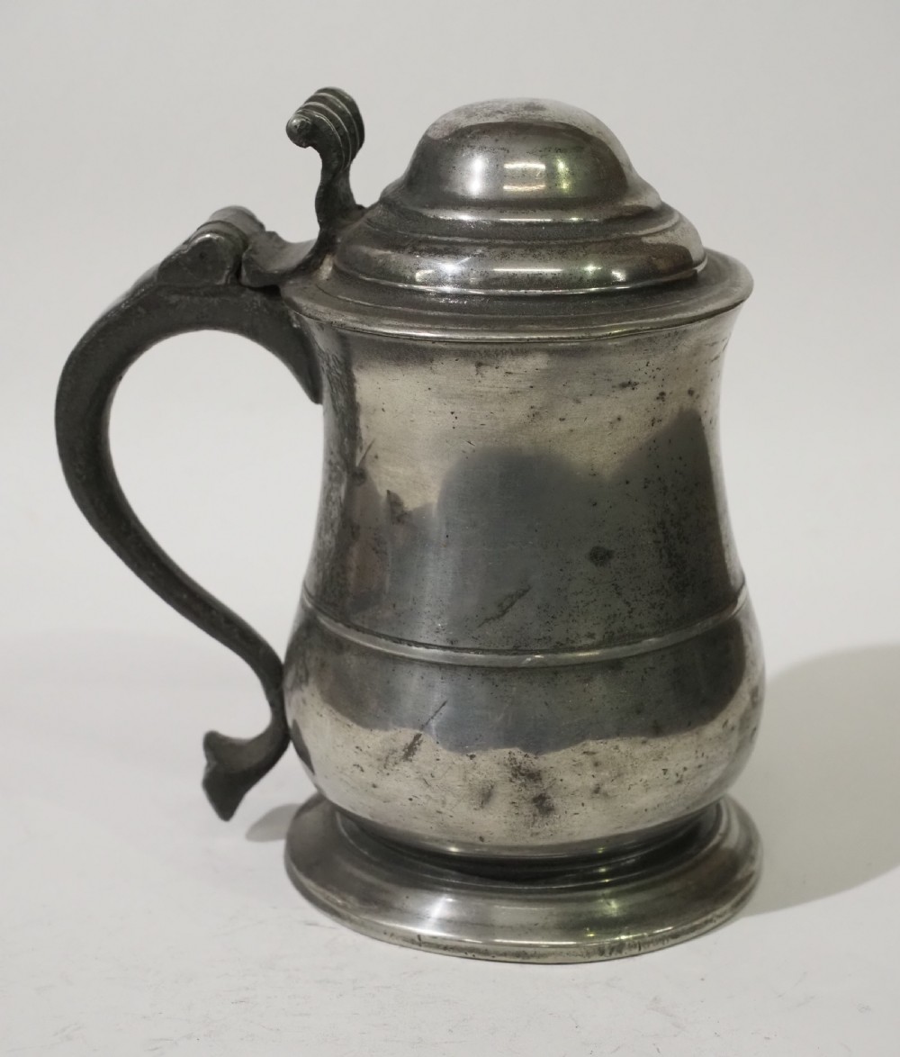 An 18th-century pewter pint tankard featuring a hinged dome lid, a curved scroll handle with a thumbpiece, and a moulded central band on a circular footed base. View II