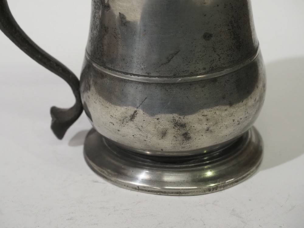 An 18th-century pewter pint tankard featuring a hinged dome lid, a curved scroll handle with a thumbpiece, and a moulded central band on a circular footed base. View IV