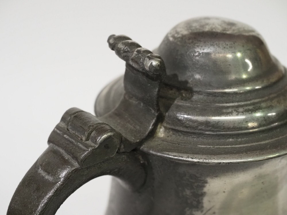 An 18th-century pewter pint tankard featuring a hinged dome lid, a curved scroll handle with a thumbpiece, and a moulded central band on a circular footed base. View V