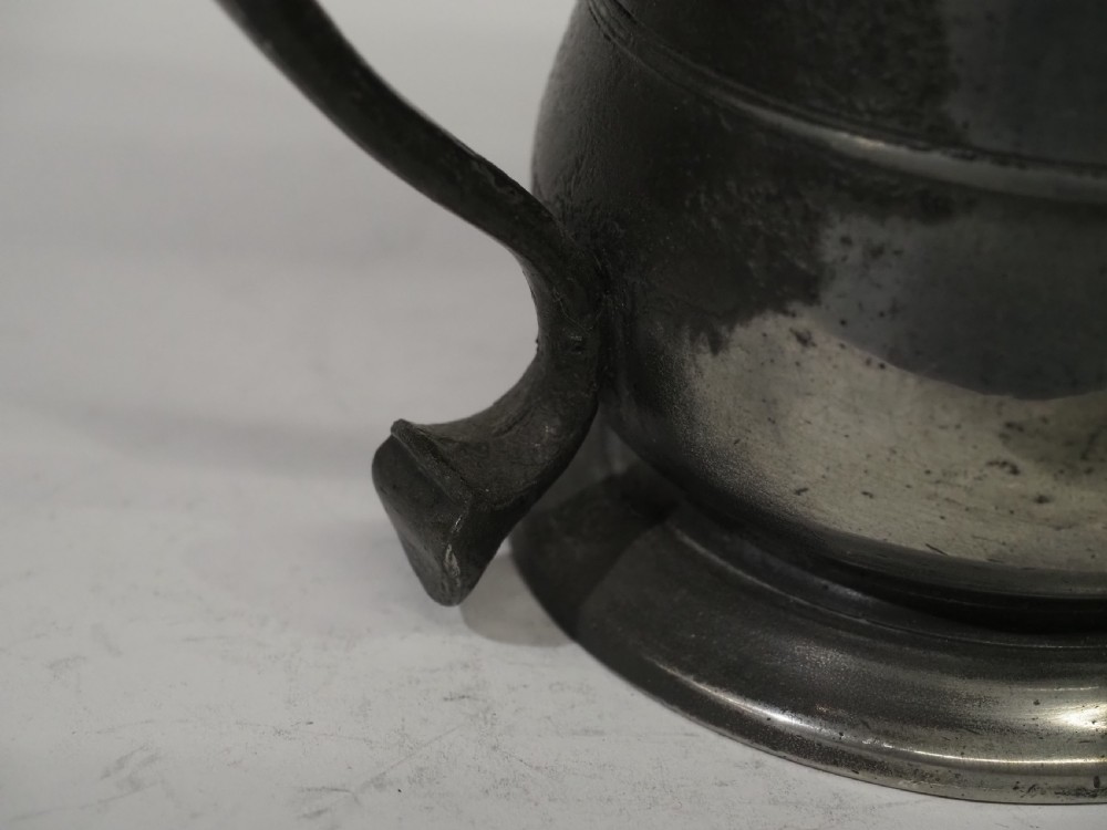 An 18th-century pewter pint tankard featuring a hinged dome lid, a curved scroll handle with a thumbpiece, and a moulded central band on a circular footed base. View VI
