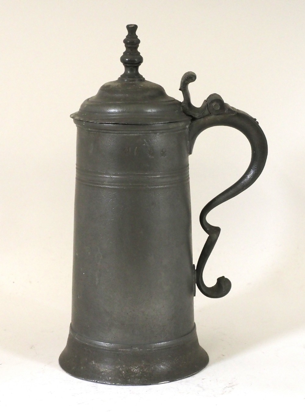 A dark grey early 18th-century English pewter spire flagon with a hinged lid, decorative spire finial, curved handle, and flared base.