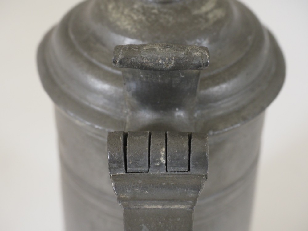 A dark grey early 18th-century English pewter spire flagon with a hinged lid, decorative spire finial, curved handle, and flared base. View V