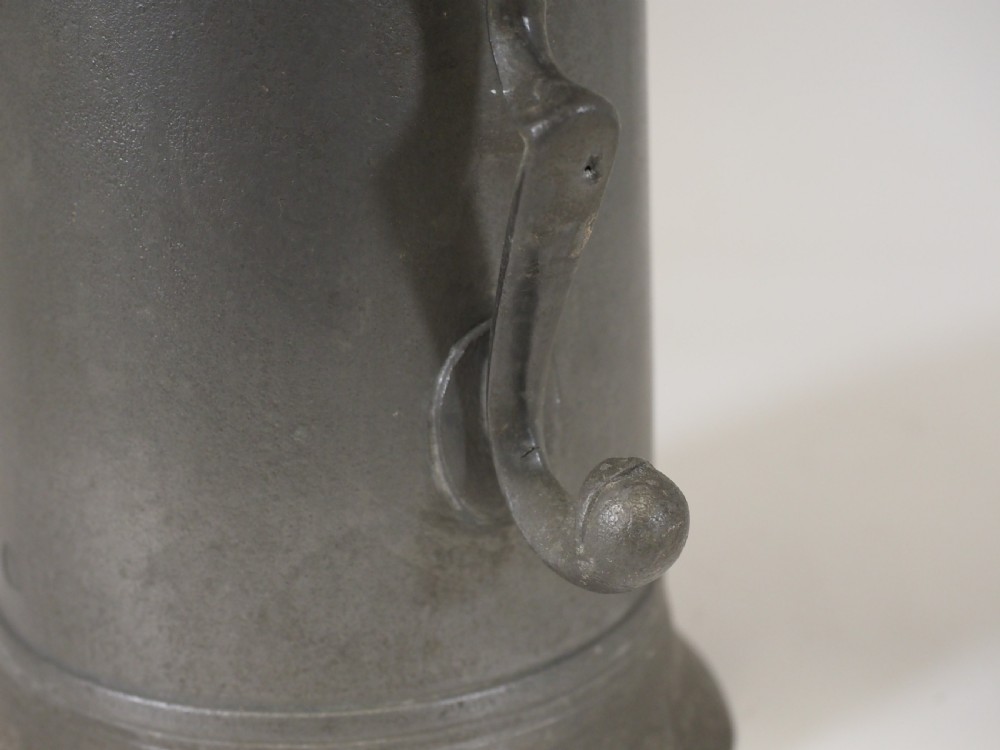 A dark grey early 18th-century English pewter spire flagon with a hinged lid, decorative spire finial, curved handle, and flared base. View VI