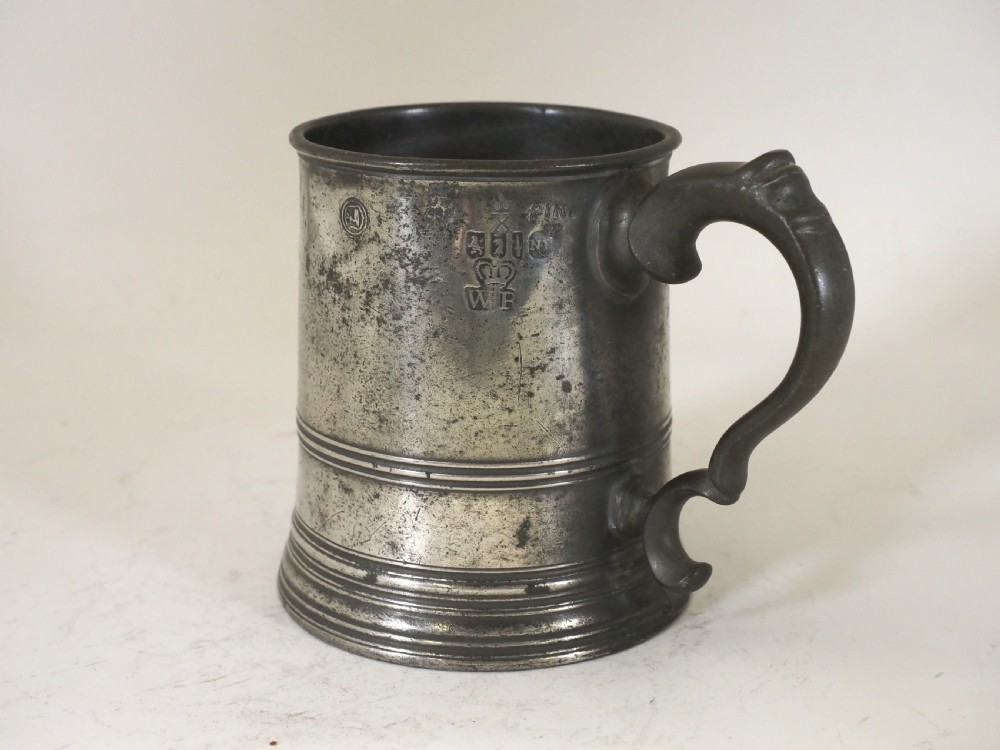 A cylindrical pewter pint measure featuring a curved handle, horizontal molded bands around the base, and stamped verification marks near the upper rim.