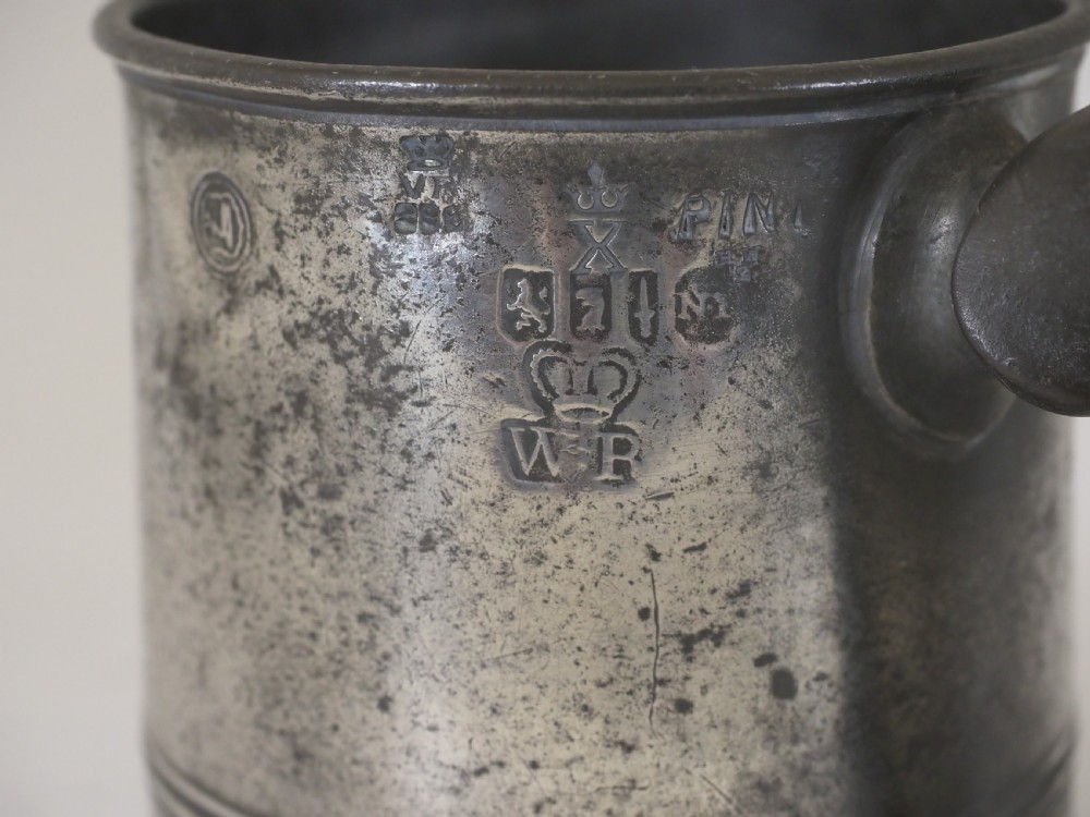 A cylindrical pewter pint measure featuring a curved handle, horizontal molded bands around the base, and stamped verification marks near the upper rim. View II
