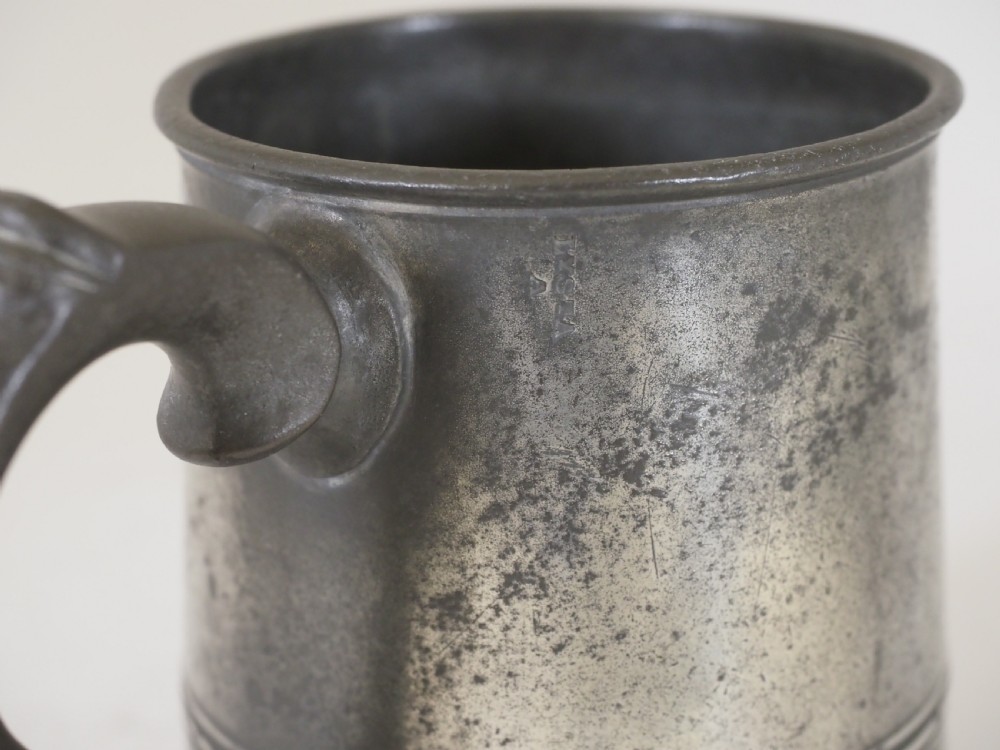 A cylindrical pewter pint measure featuring a curved handle, horizontal molded bands around the base, and stamped verification marks near the upper rim. View V