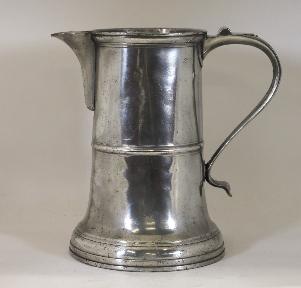 An 18th-century English pewter ale jug featuring a flared base, central girdle moulding, single poured spout, and curved handle.