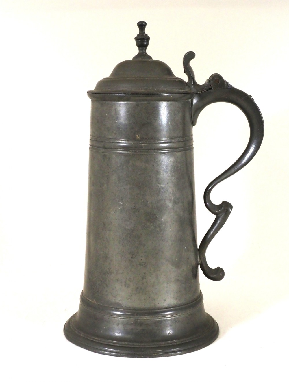 An eighteenth-century dark grey pewter spire flagon featuring a tapered cylindrical body, domed lid with spire finial, double-scroll handle, and moulded spreading base.