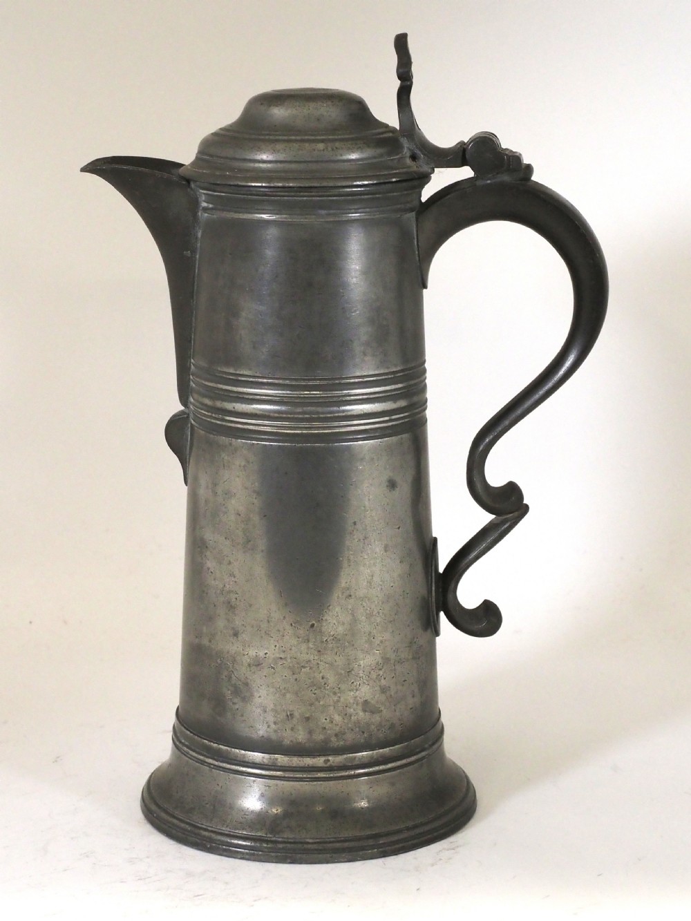 An antique late 18th-century spouted pewter spire flagon featuring a double-scroll handle, domed lid, and flared circular base.