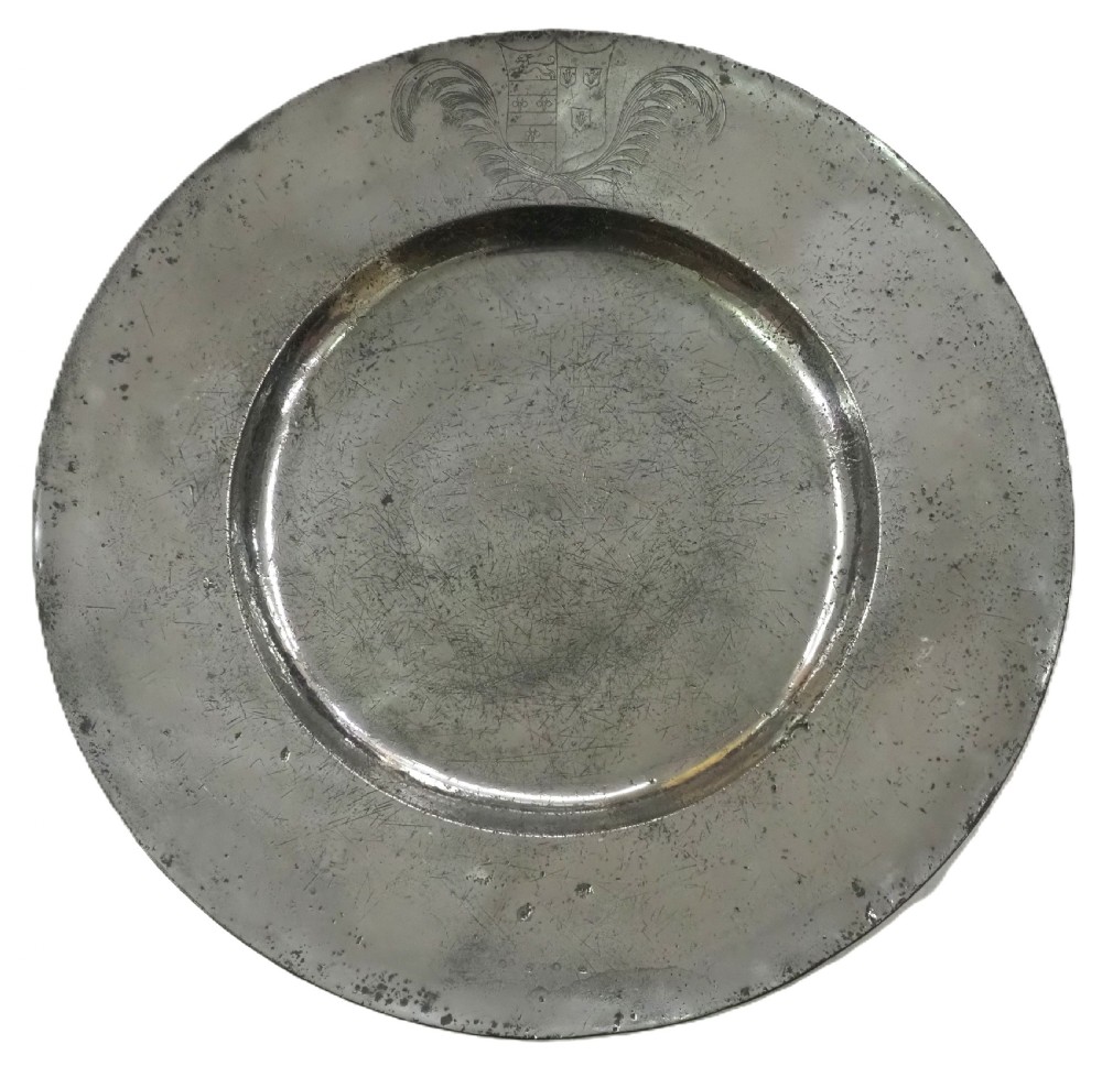 A circular seventeenth-century pewter broad rim charger featuring an engraved coat of arms flanked by feathered plumes on the upper rim.