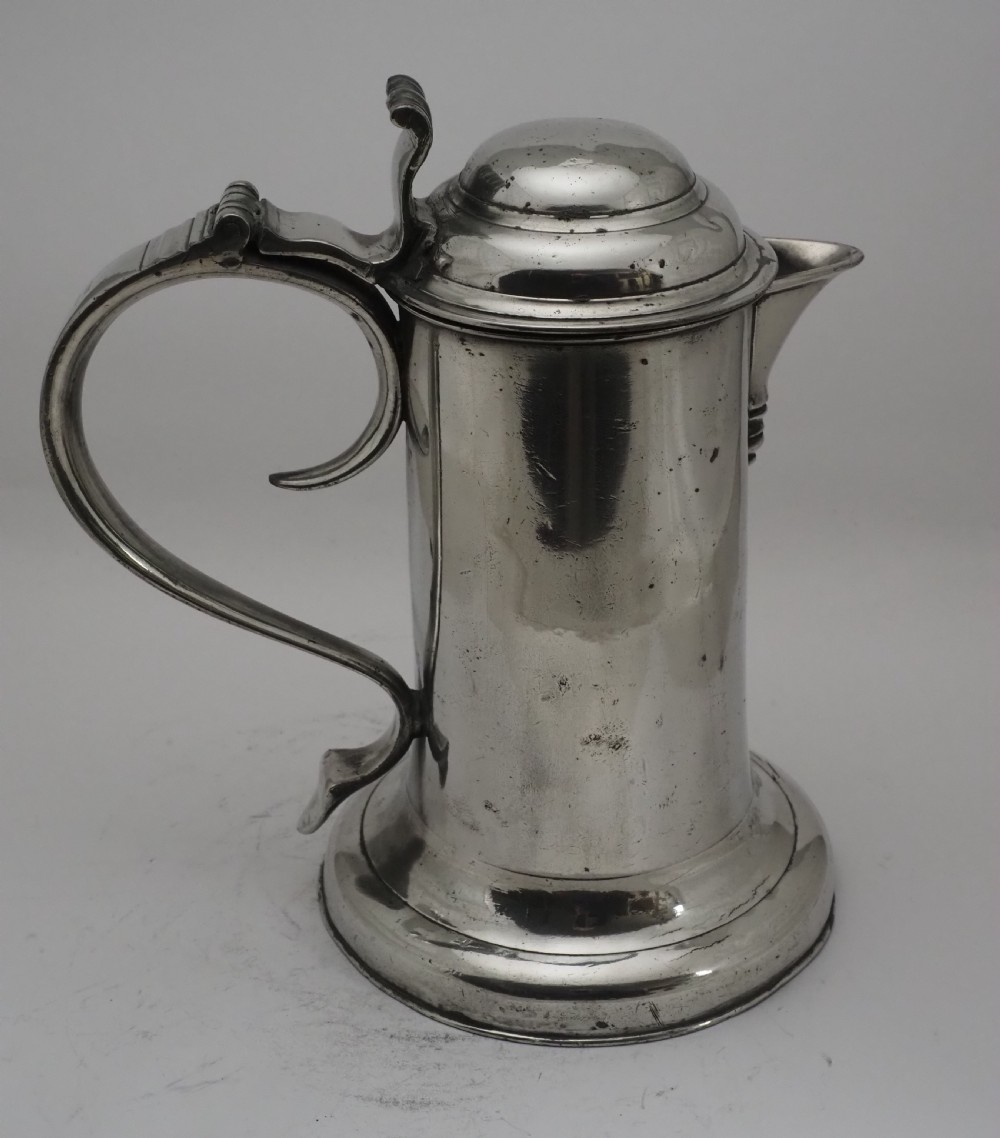 An 18th-century Irish pewter church flagon with a domed hinged lid, thumbpiece, curved handle, and broad flared base. View XVI