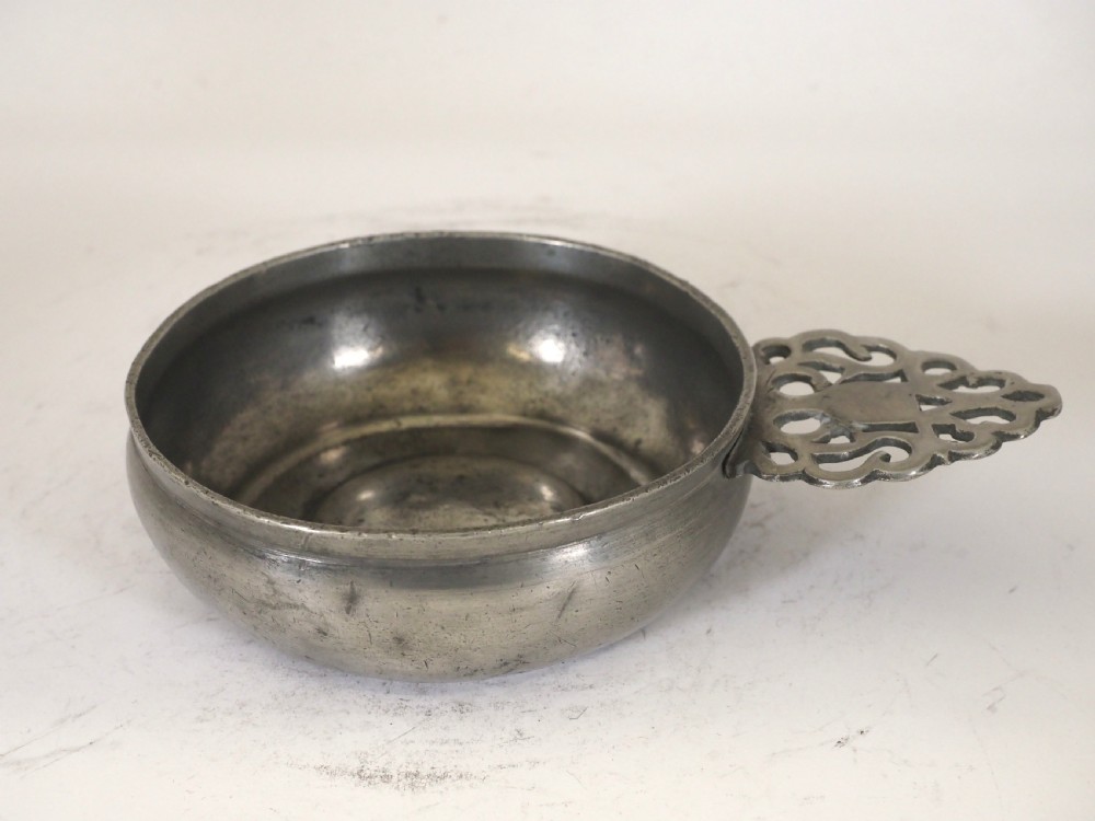 An antique silver-grey pewter porringer bowl featuring a single ornate pierced scrollwork ear handle.