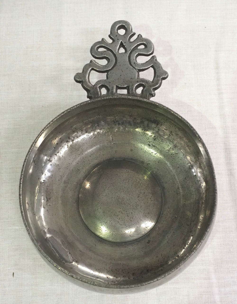 An antique circular pewter porringer bowl featuring a textured interior basin and a decorative pierced scrollwork handle. View II