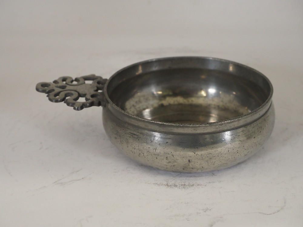 An antique circular pewter porringer bowl featuring a textured interior basin and a decorative pierced scrollwork handle. View IV