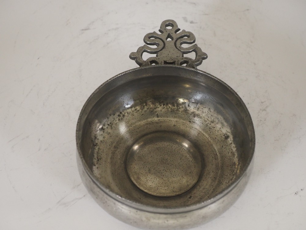 An antique circular pewter porringer bowl featuring a textured interior basin and a decorative pierced scrollwork handle. View V