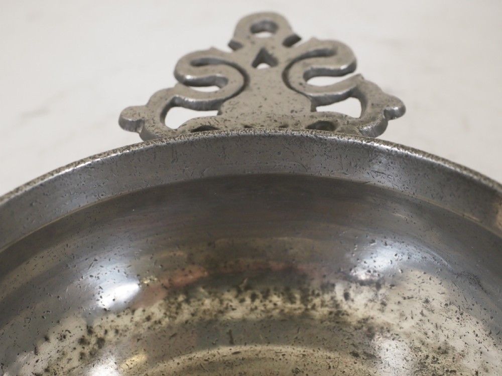 An antique circular pewter porringer bowl featuring a textured interior basin and a decorative pierced scrollwork handle. View VII