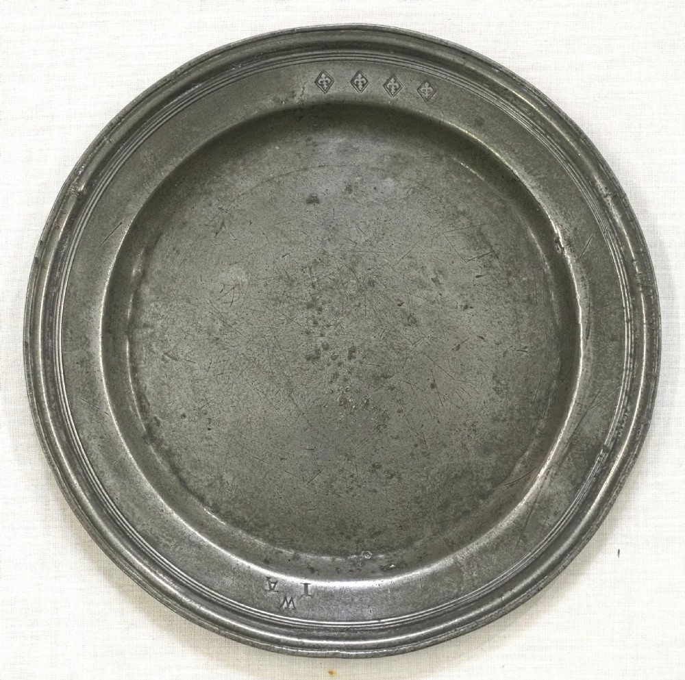 A circular 17th-century English pewter plate displaying a multiple reeded rim with stamped hallmarks near the top edge and initial stamps near the bottom.