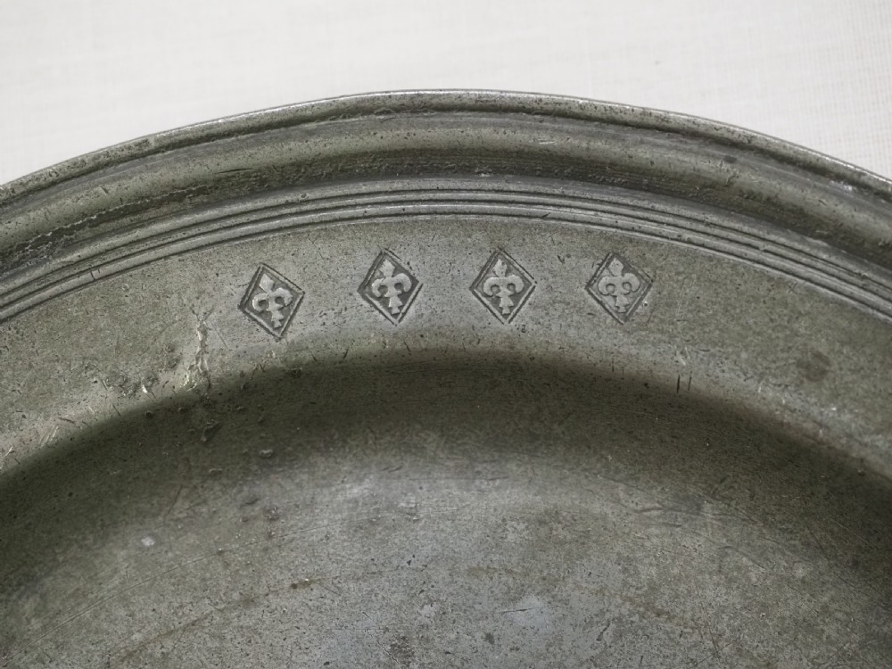 A circular 17th-century English pewter plate displaying a multiple reeded rim with stamped hallmarks near the top edge and initial stamps near the bottom. View II
