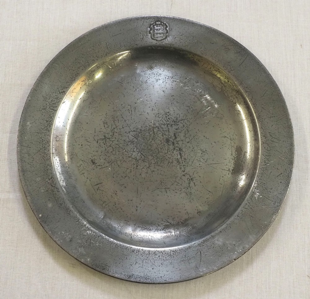 A circular 18th-century English pewter plate with a broad rim featuring a small stamped coat of arms at the top.