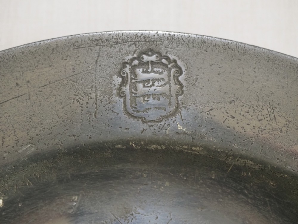 A circular 18th-century English pewter plate with a broad rim featuring a small stamped coat of arms at the top. View II