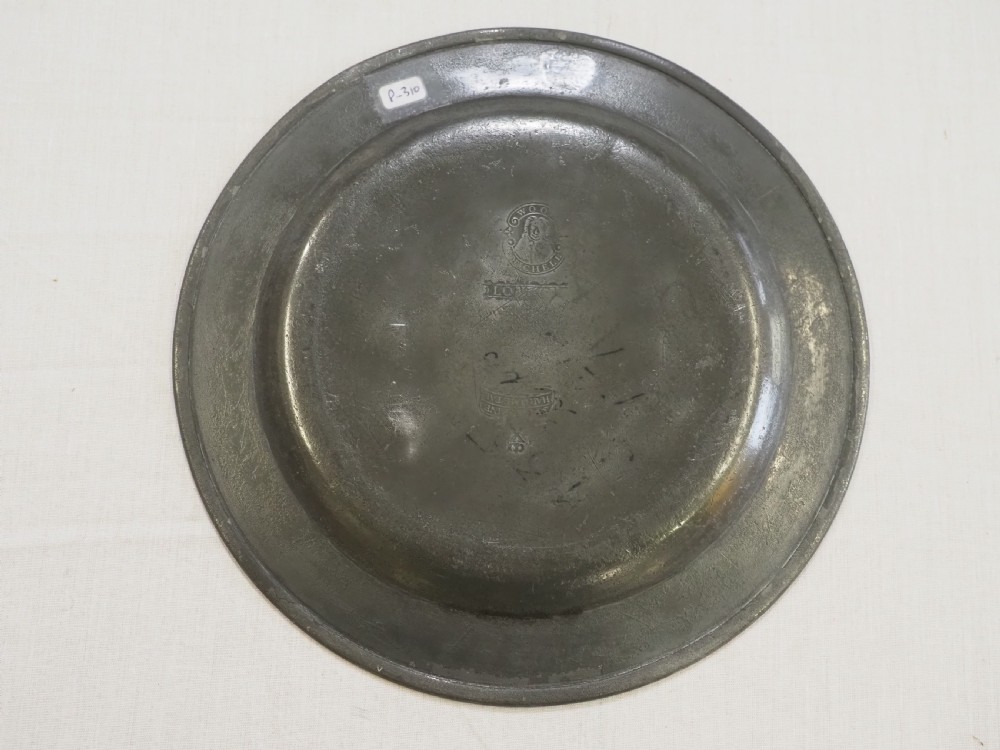 A circular 18th-century English pewter plate with a broad rim featuring a small stamped coat of arms at the top. View IV