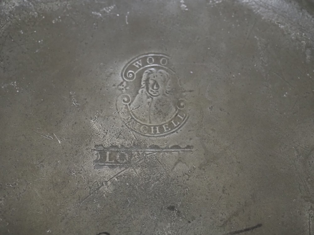 A circular 18th-century English pewter plate with a broad rim featuring a small stamped coat of arms at the top. View V