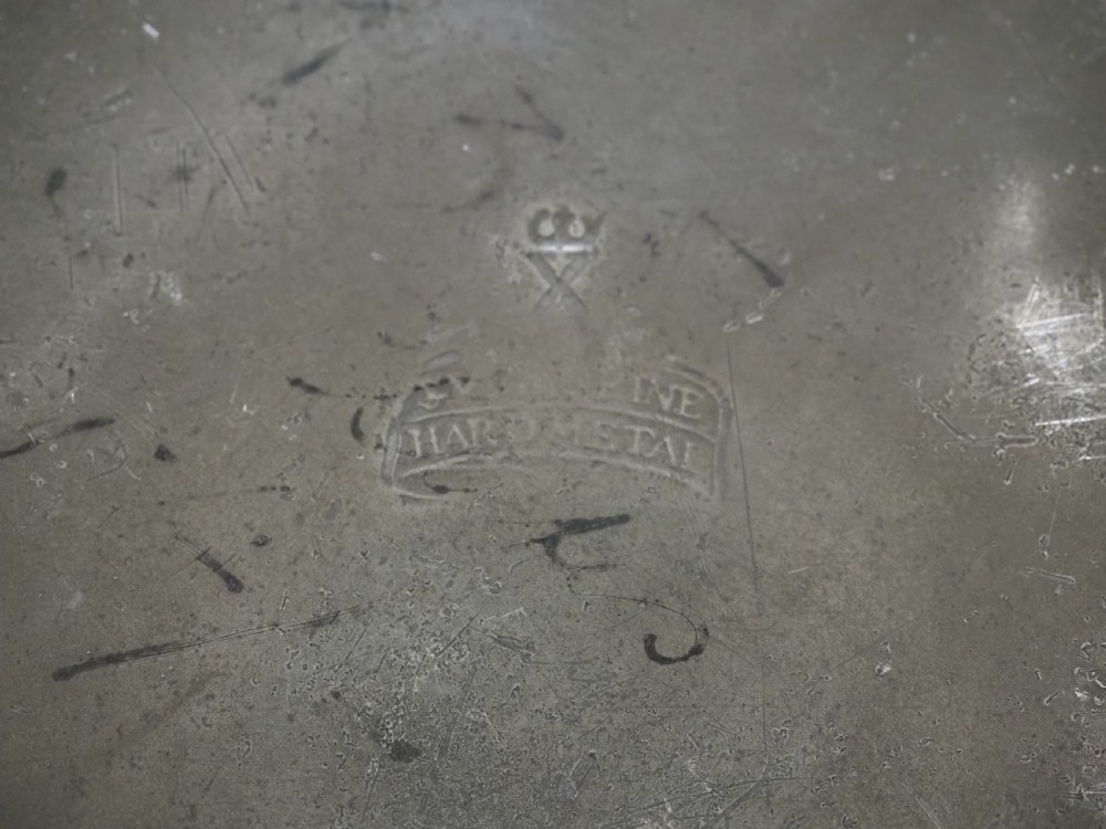 A circular 18th-century English pewter plate with a broad rim featuring a small stamped coat of arms at the top. View VI