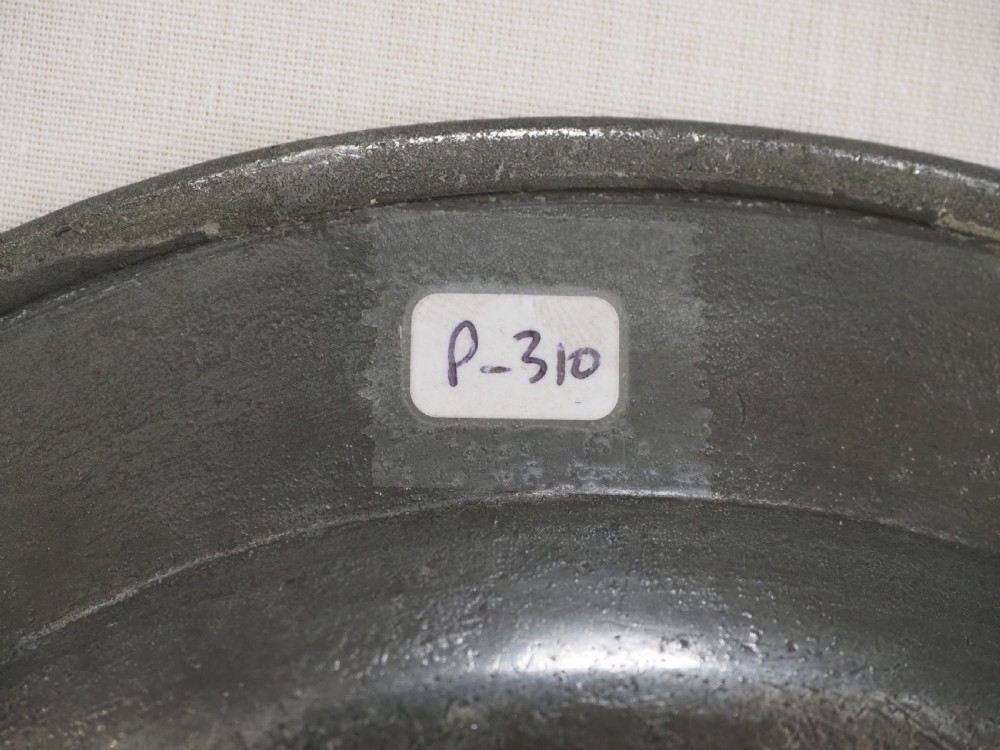A circular 18th-century English pewter plate with a broad rim featuring a small stamped coat of arms at the top. View VII