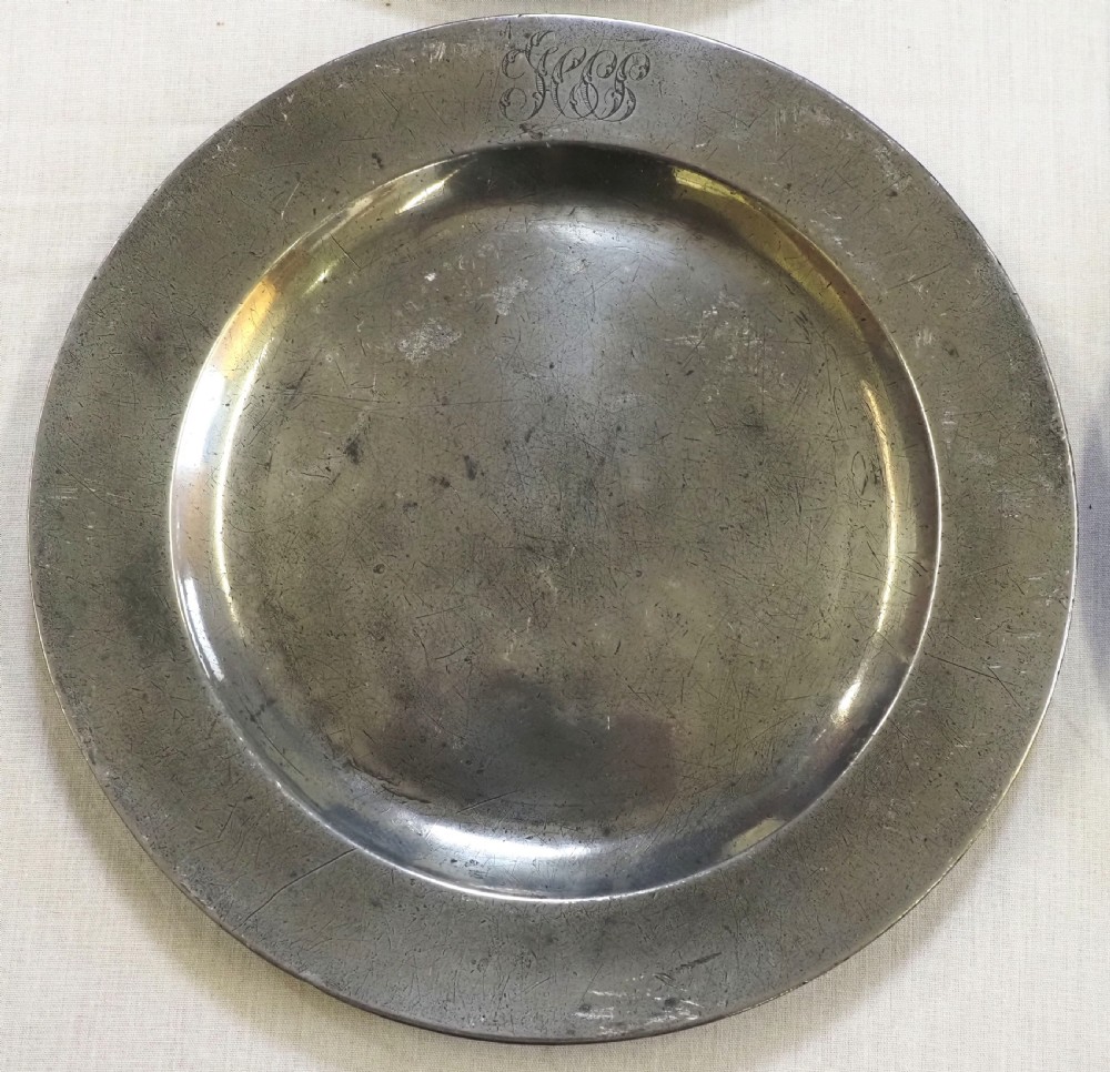 A set of four circular antique pewter plates featuring engraved monograms along the upper rims and a naturally aged grey surface. View IV
