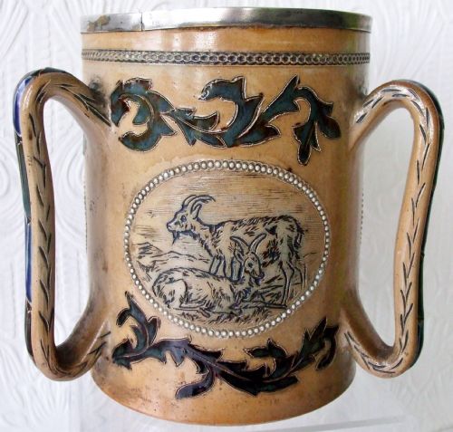 fig 1. An antique tan stoneware tyg featuring three large handles, a silver-mounted rim, and an incised central medallion depicting a donkey surrounded by dark foliage scrollwork.