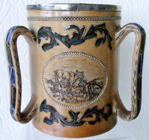 fig 2. An antique tan stoneware tyg featuring three large handles, a silver-mounted rim, and an incised central medallion depicting a donkey surrounded by dark foliage scrollwork.